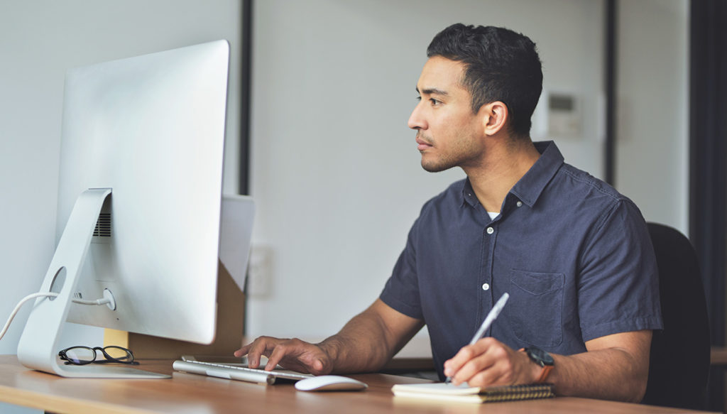 man looking at computer screen and writing down notes